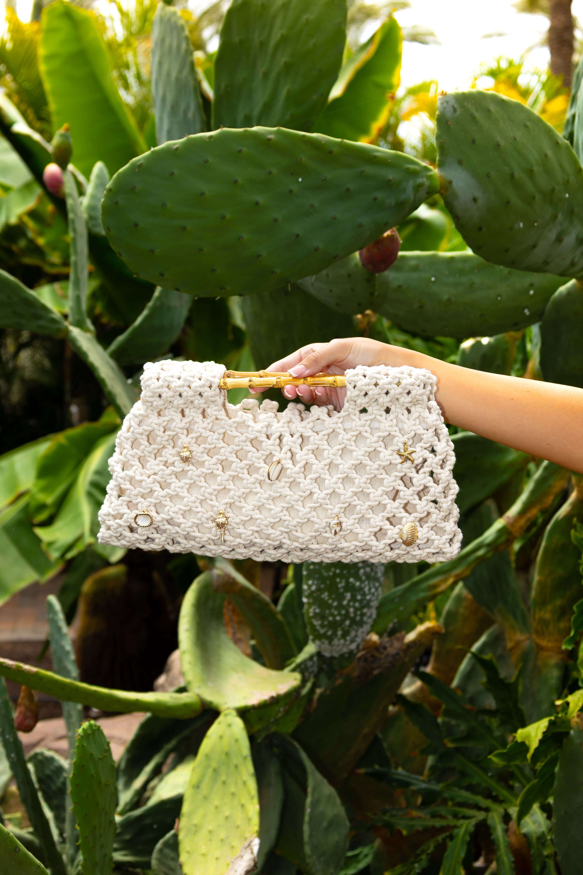 Hand holding a cream white crochet beach bag with gold shell charms in front of a cactus