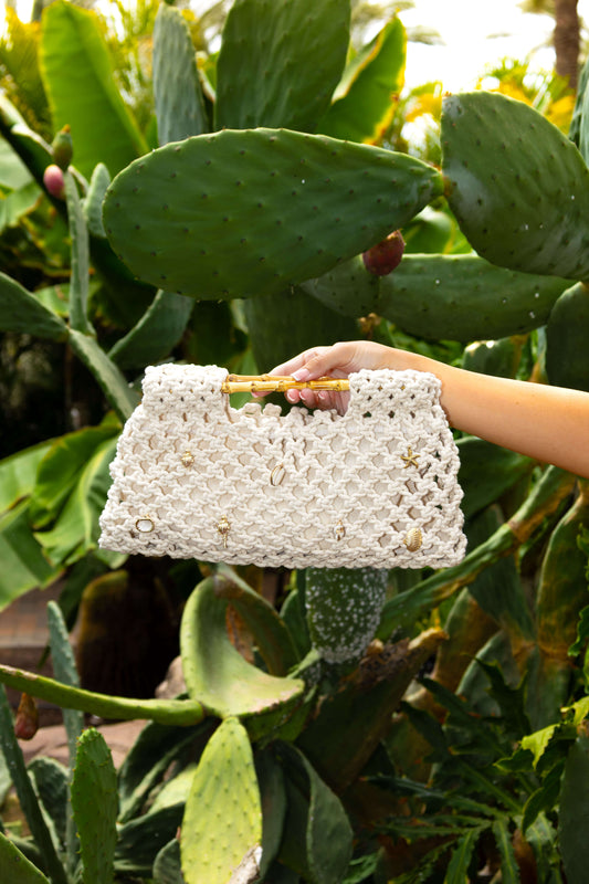 Hand holding a cream white crochet beach bag with gold shell charms in front of a cactus