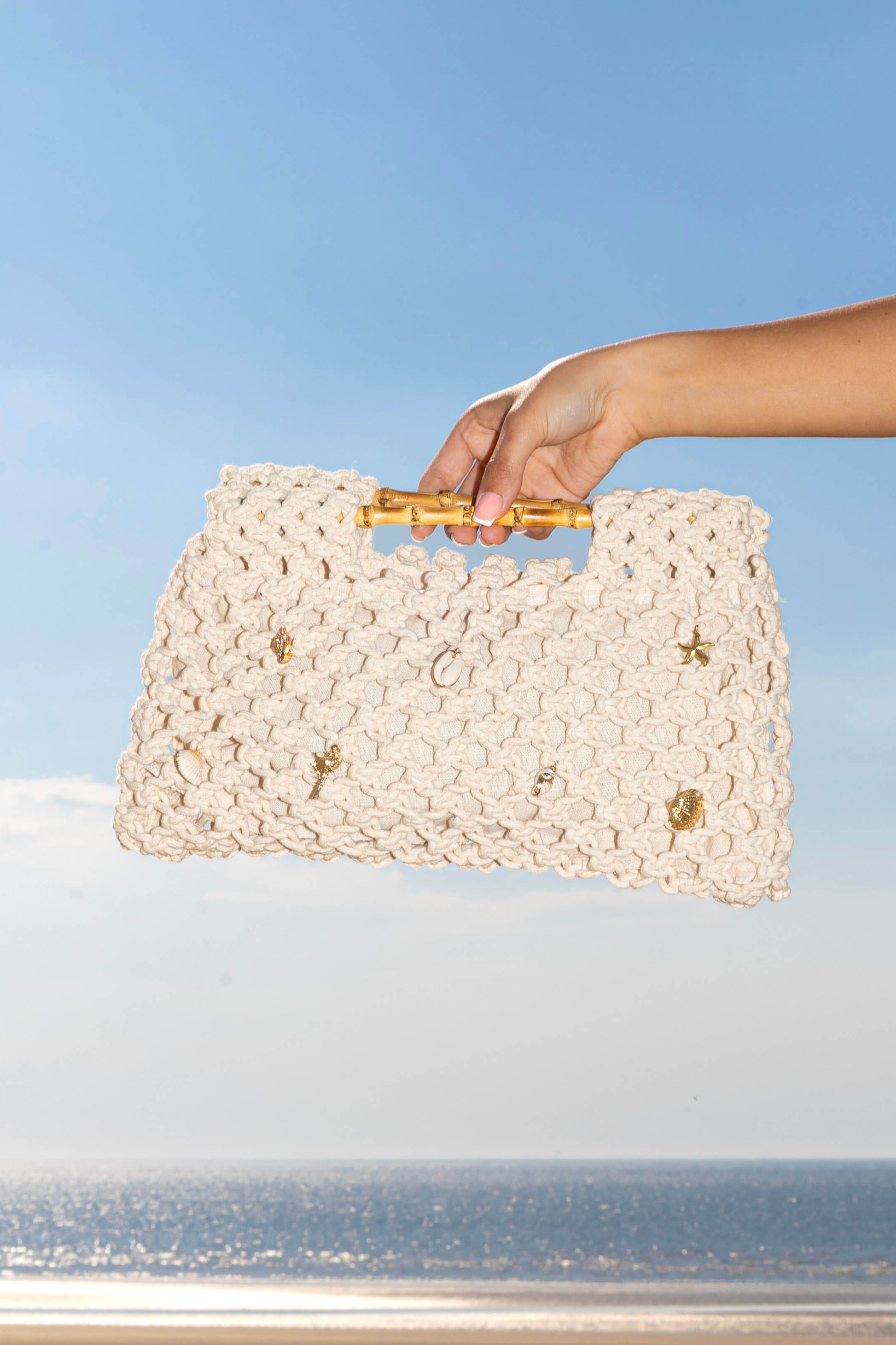 Hand holding a woven crochet cream white clutch bag against a beach background