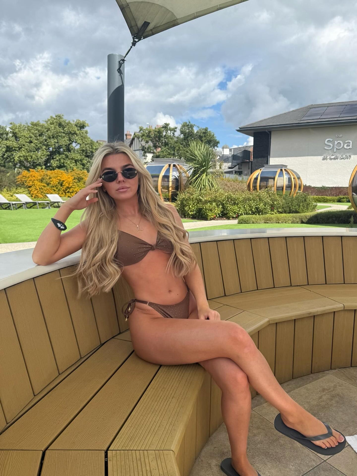 Woman in a brown by opulence bikini sitting on a wooden bench outdoors with a spa in the background.