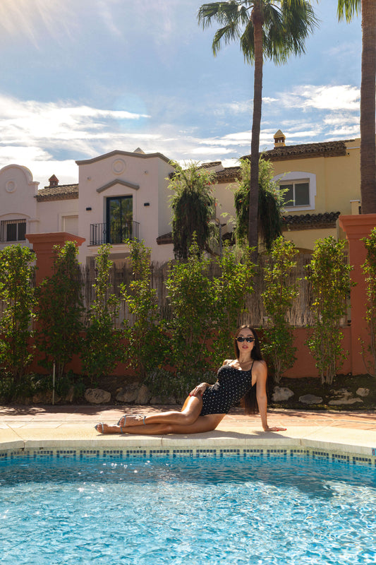 A woman wearing a black diamante by Opulence swimsuit and sunglasses relaxes by a swimming pool with a resort-style building, palm trees, and a wooden fence in the background.