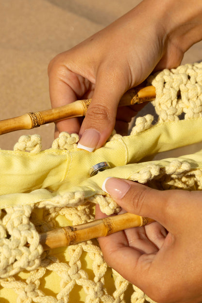Close-up of hands holding a woven yellow beach bag with bamboo handles against a blurred sand background.