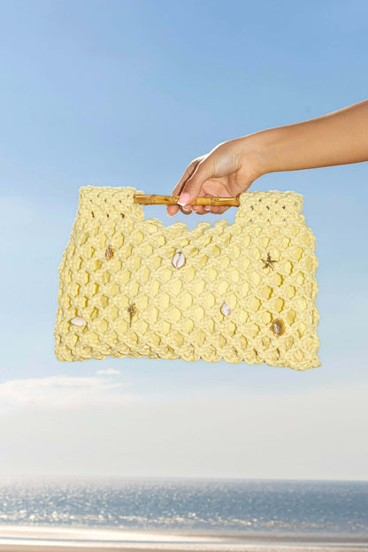 Hand holding a yellow crochet clutch beach bag with a blue sky and ocean background