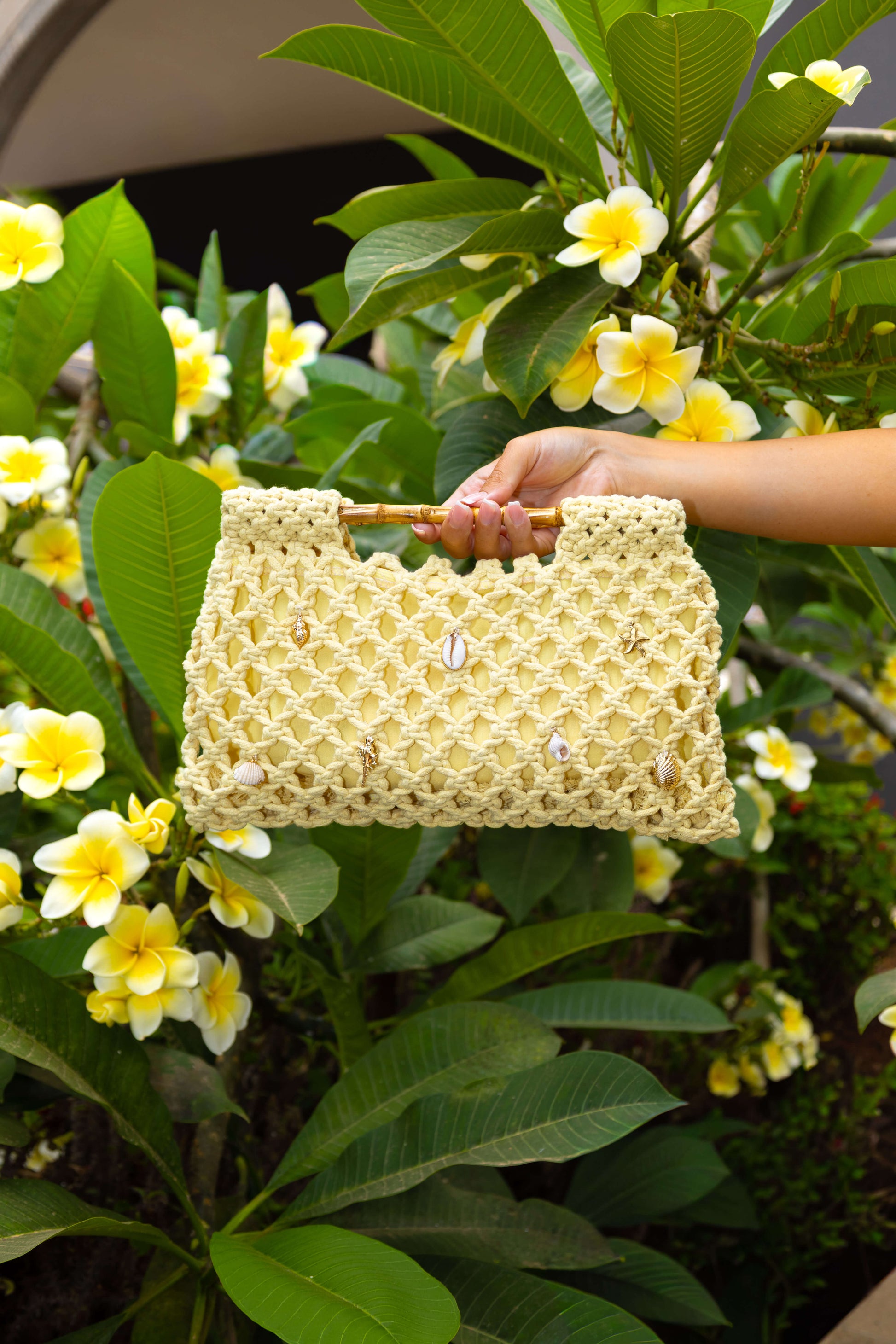Hand holding a yellow crochet beach bag featuring bamboo handles, real shells and gold charms 