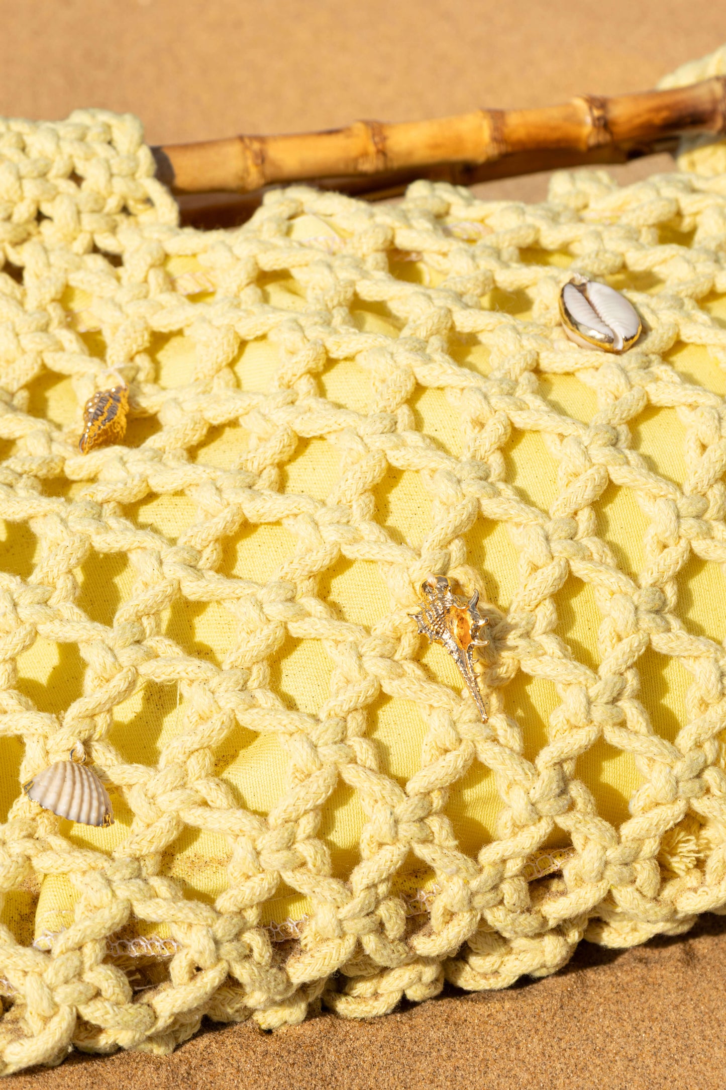 Close-up of a yellow woven crochet beach bag with shells and gold charms on a sand background