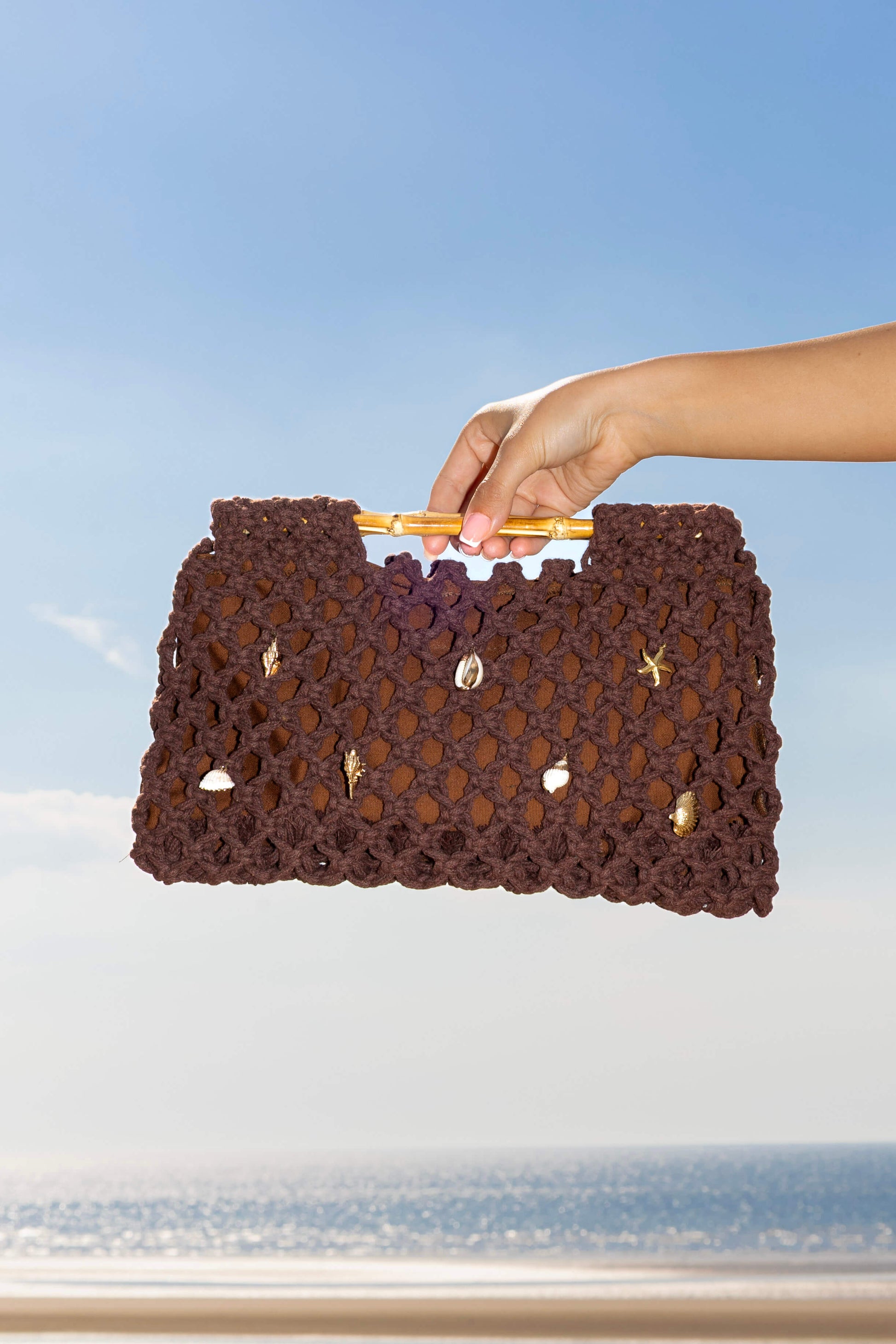 Hand holding a brown crochet clutch beach bag against a blue sky and ocean background