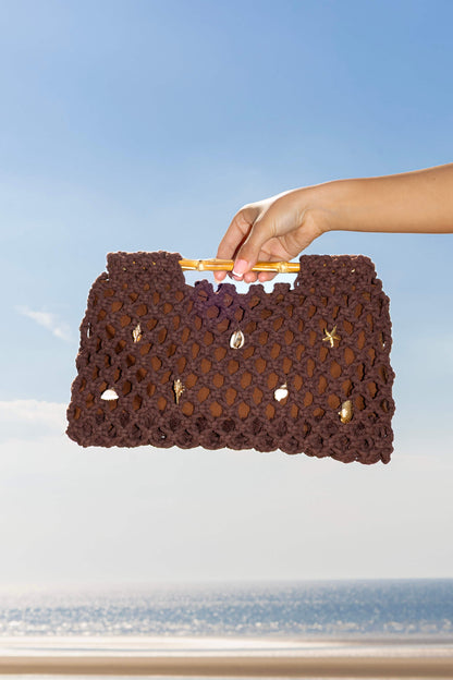 Hand holding a brown crochet clutch beach bag against a blue sky and ocean background