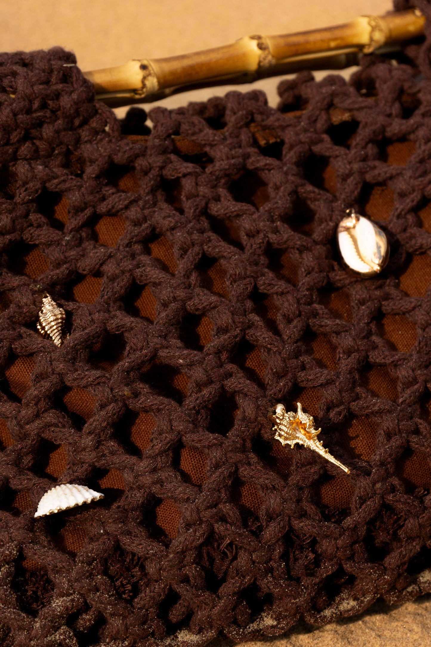 Close-up of a brown crochet beach bag with gold shell charms on sand