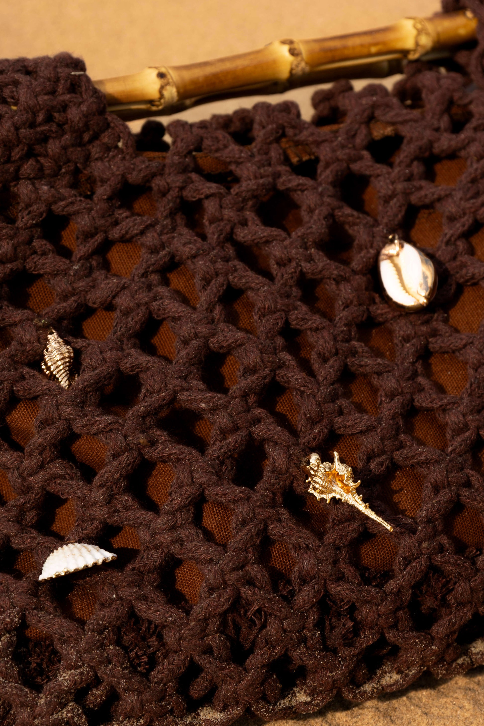Close-up of a brown crochet beach bag with gold shell charms on sand
