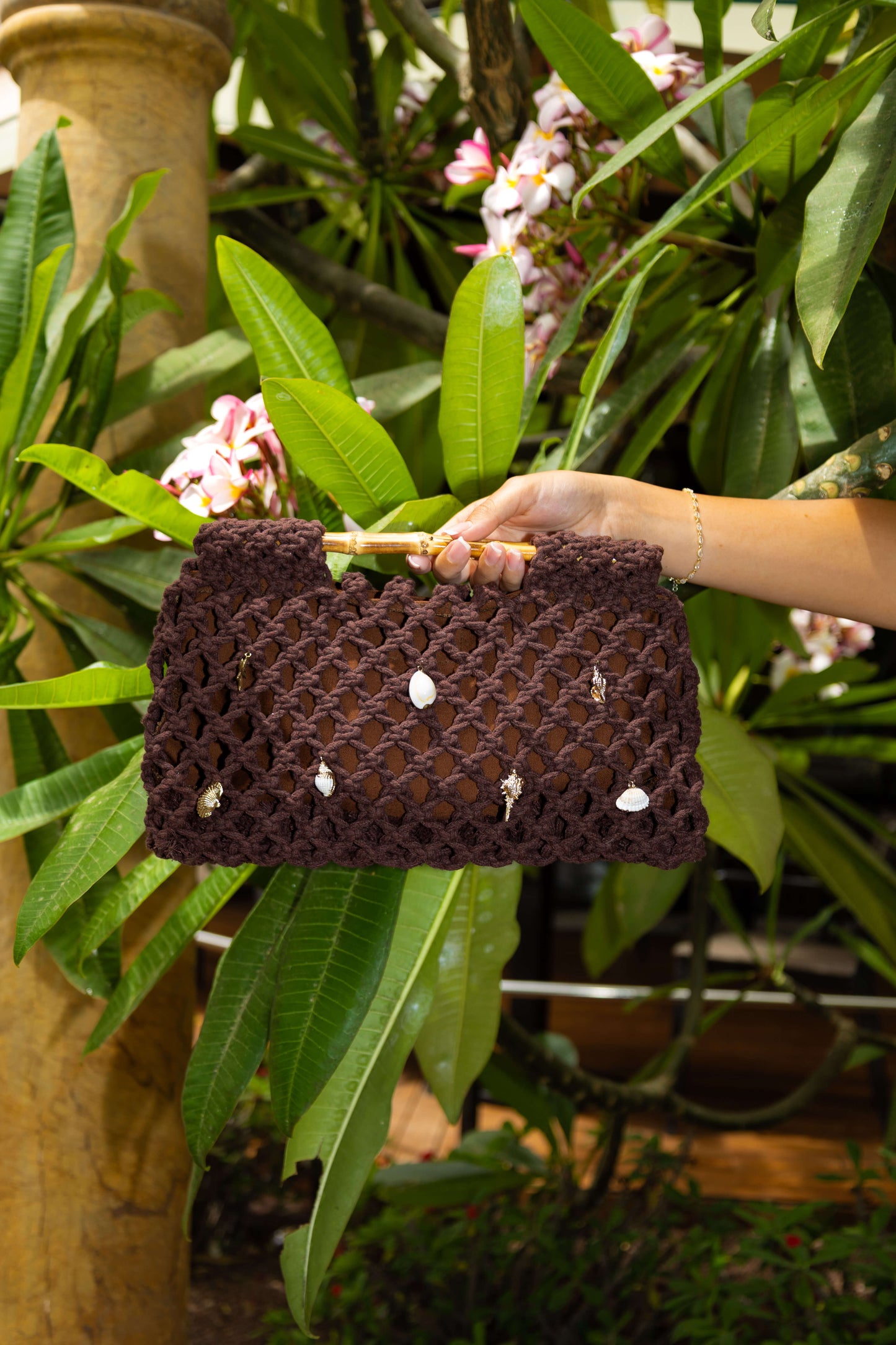 Brown woven crochet beach bag with charms held by a person with green tropical leaves in the background
